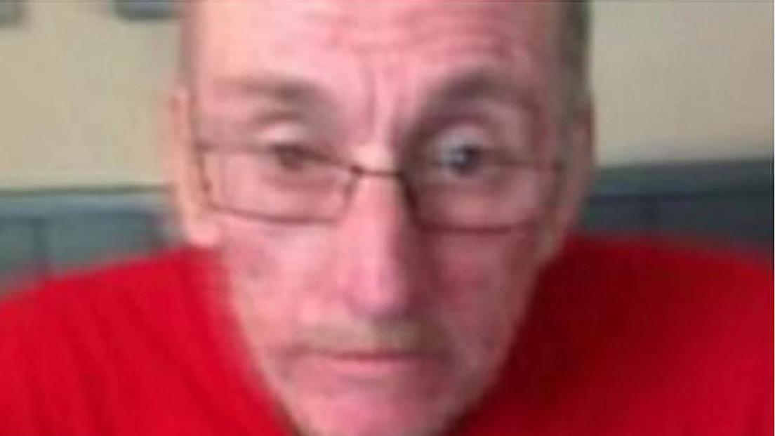 Tony Nicholls is seen in a blurry photograph. He is wearing glasses and a red jumper and is looking serious.