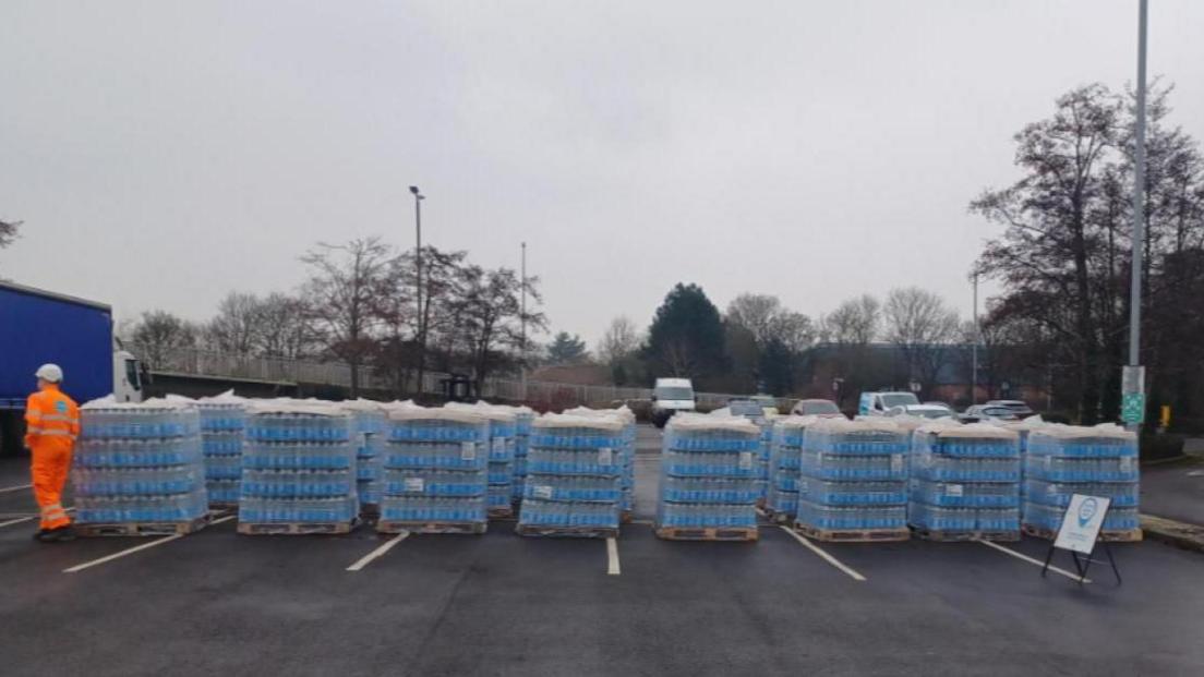 Around 20 wooden pallet crates stacked high with bottled of water, wrapped and sealed in clear plastic. They are sitting in the middle of a car park on a grey and dreary day. On the left there is a Thames Water employee dressed in full hi-vis orange with a white plastic helmet.