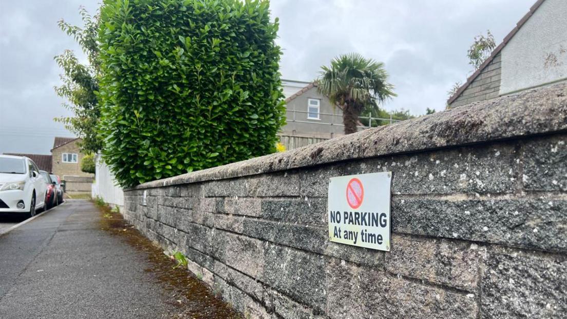 A sign on a low garden wall that says 'No parking at any time'. On the left of the picture you can see cars parked alongside the road.