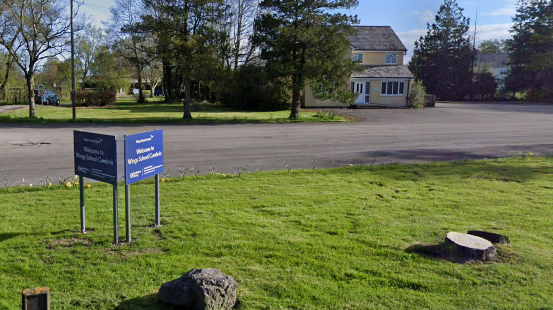 Blue signage on a small  grass section reads 'Wings School Cumbria'. There is a large tarmaced driveway with trees and a large residential property. 