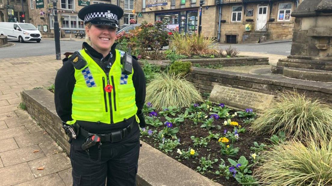 A uniformed police officer stands next to a public flowerbed. A poppy is pinned to the middle of her luminous waistcoat.