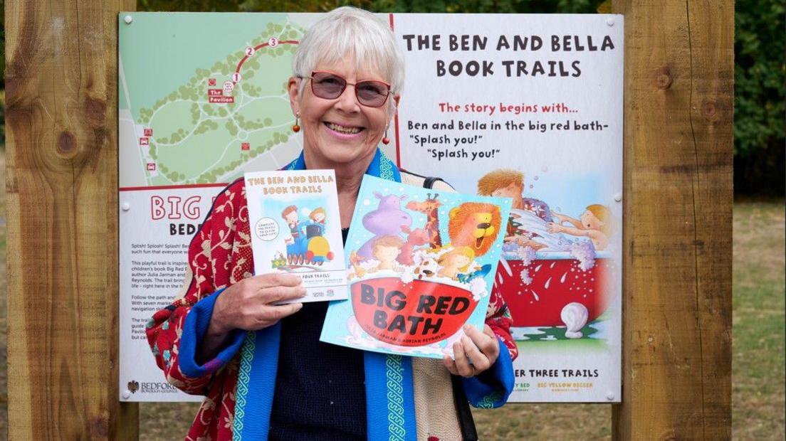 Julia Jarman, holding two story books, by a sign for a book trail. She is smiling, looking at the camera, she has on glasses, earrings, and is wearing a colourful coat.