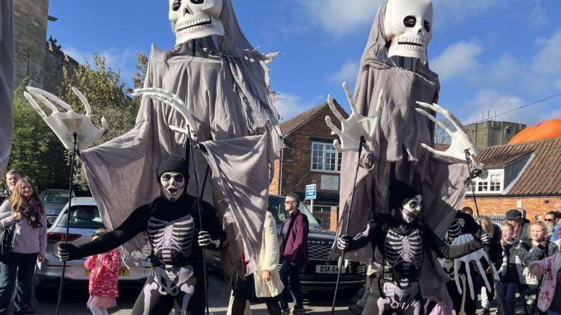 Two people dressed as black and white skeletons. They have larger skeleton figures attached to them. They have skeleton face paint on and have their mouths open. They are dancing and members of the public are watching. 