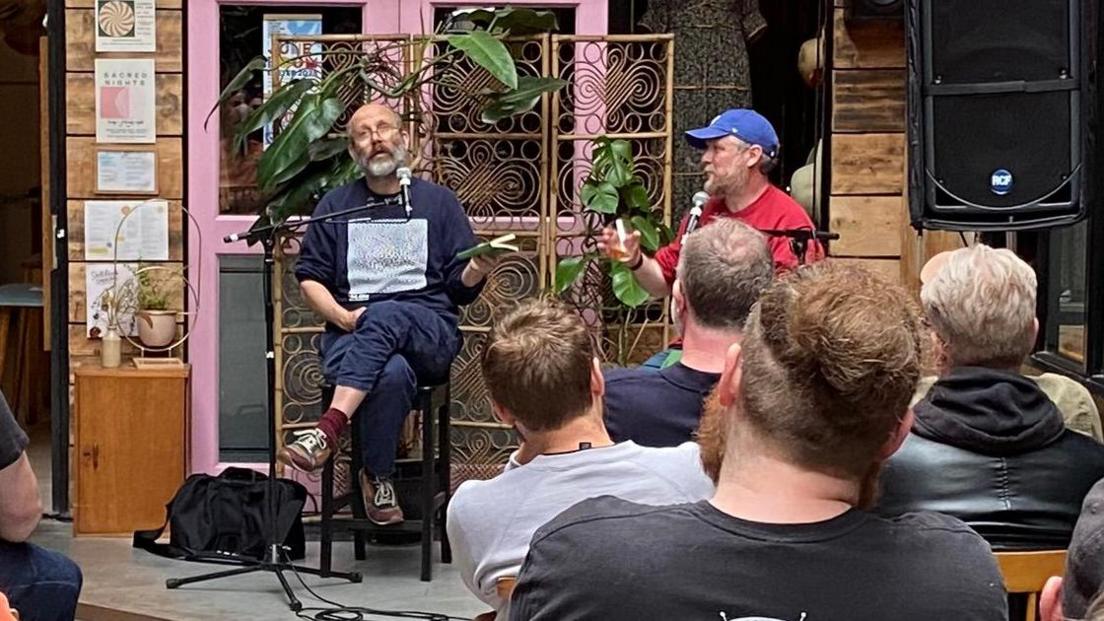 Daniel Kitson, a bald man with a beard, sat besides Tim Key, a man with a beard wearing a blue cap, on stage