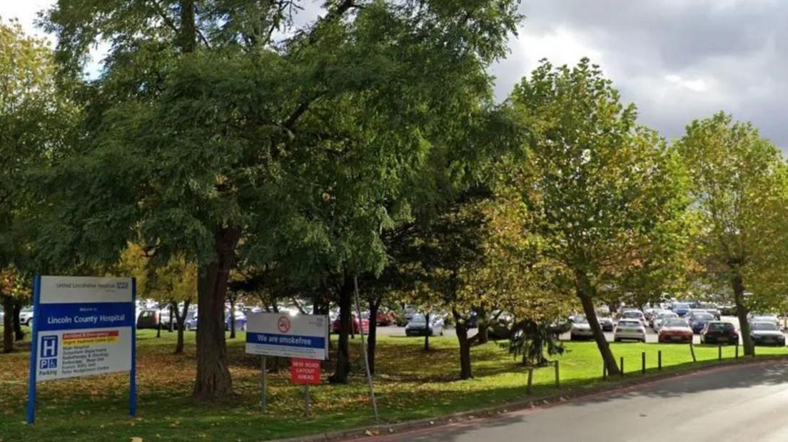 The entrance to a car park at Lincoln County Hospital. A blue and white sign on the grass at the side of the road surrounded by several trees. Several cars are in a car park in the distance.