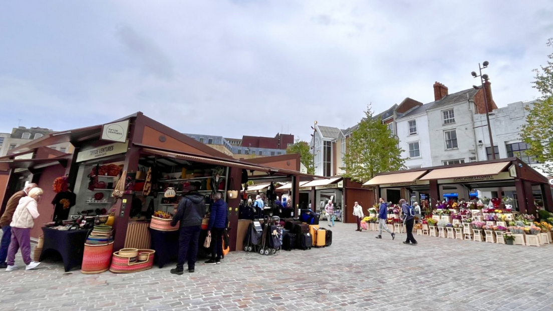 Brown-coloured market stalls selling woven baskets, luggage and flowers while people walk around on cobbled paving 