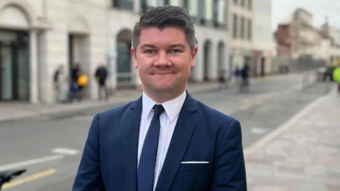 A picture of Sam smiling at the camera. He has black hair and is wearing a white suit shirt, navy tie and a navy blazer.
