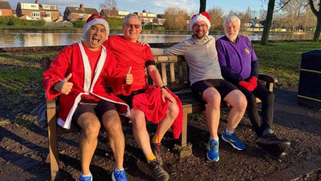 Four people dressed in running gear sat on a bench smiling at the camera. Two men are wearing Santa hats and one is wearing a Santa jacket.