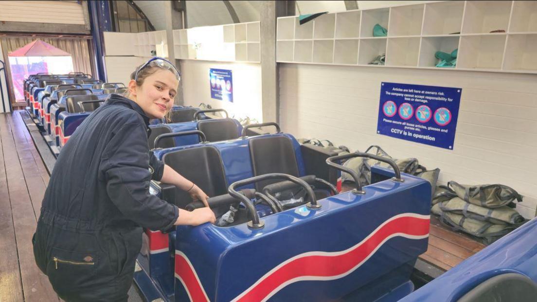 A young girl wearing navy overalls, smiles at the camera as she tinkers with a rollercoaster seat 