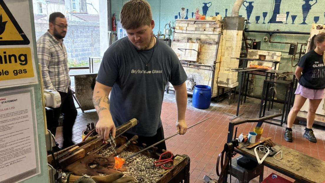 A glassblower at work with a hot, fiery-orange piece of glass at the end of a metal stick, surrounded by other tools and two other colleagues are in the background of the workshop