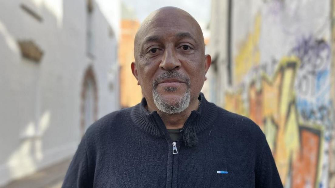 Desmond Brown in a blue zip up jumper. He is bald, middle aged with a white beard. Behind him is a street with lots of colourful graffiti on it. 