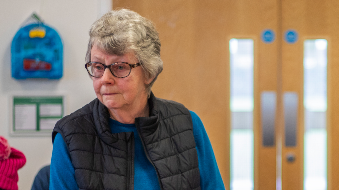 A woman with grey hair and glasses wearing a gilet over a blue jumper 