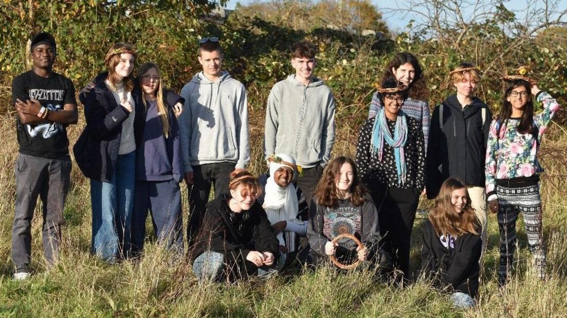 A large group of young people pose for the camera at an Avon Wildlife Trust site in Begbrook in Bristol. Many of them are wearing hoodies and some are shielding their eyes as it is a sunny day. They are standing in a meadow with long grass and a hedgerow behind them