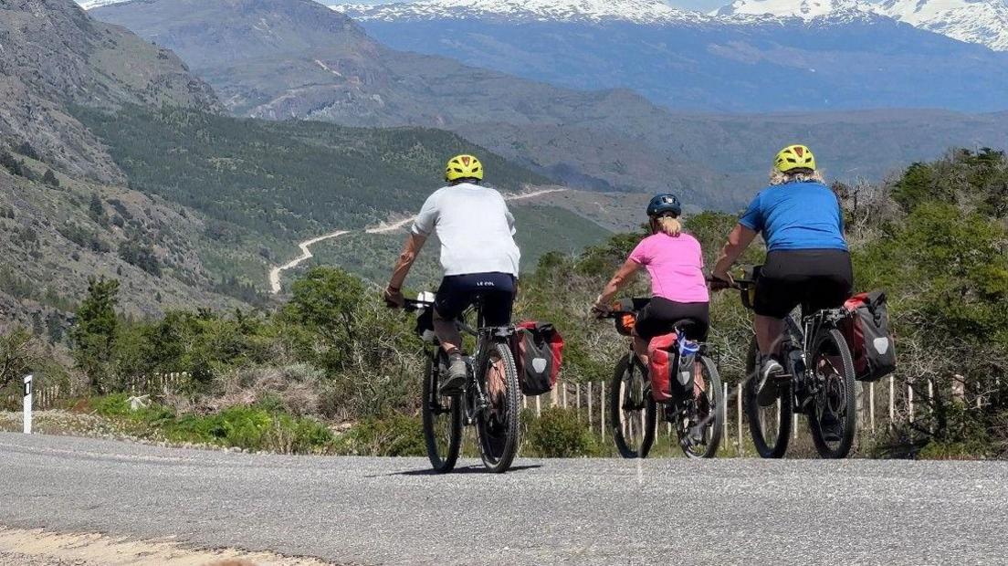 Craig, Sophie and Rona are on their bikes cycling a long a road which zig-zags up a mountainside in the distance. A mountain range rises in the distance.