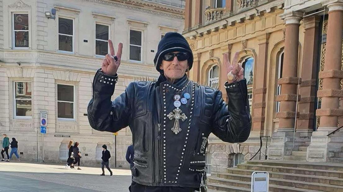 A man wearing a black leather jacket, black hat, and black glasses. He is doing a peace sign with both of his hands
