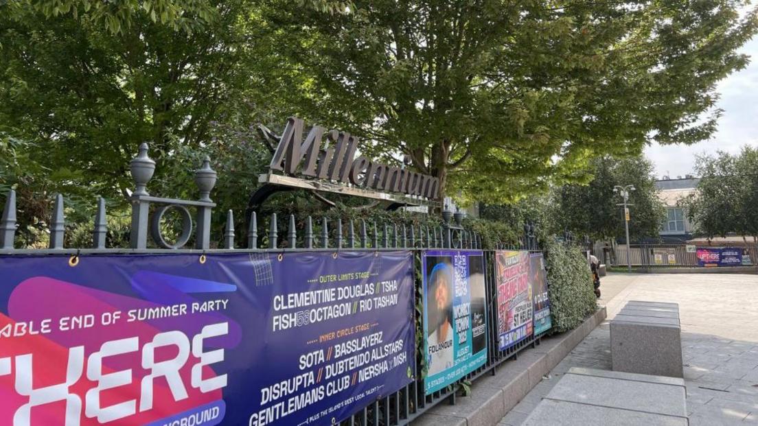 A sign for Millennium Park on the railings on the edge of the park in Jersey. Posters for events happening at the park are on the railings.