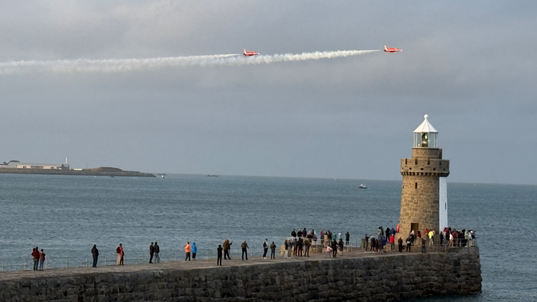 Crowds gathered on a pier in St Peter Port, Guernsey, looking into the sky out to sea, with the Red Arrows flying over in the sky.