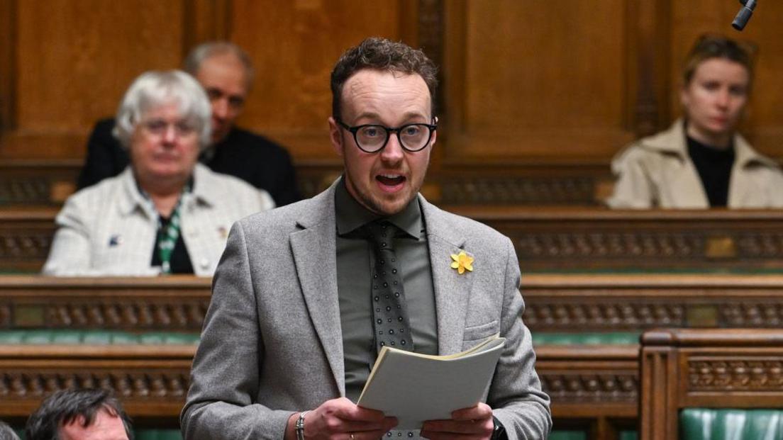 Adam Dance speaking in Parliament. He is wearing a grey jacket, shirt and tie, and has a yellow flower in his lapel