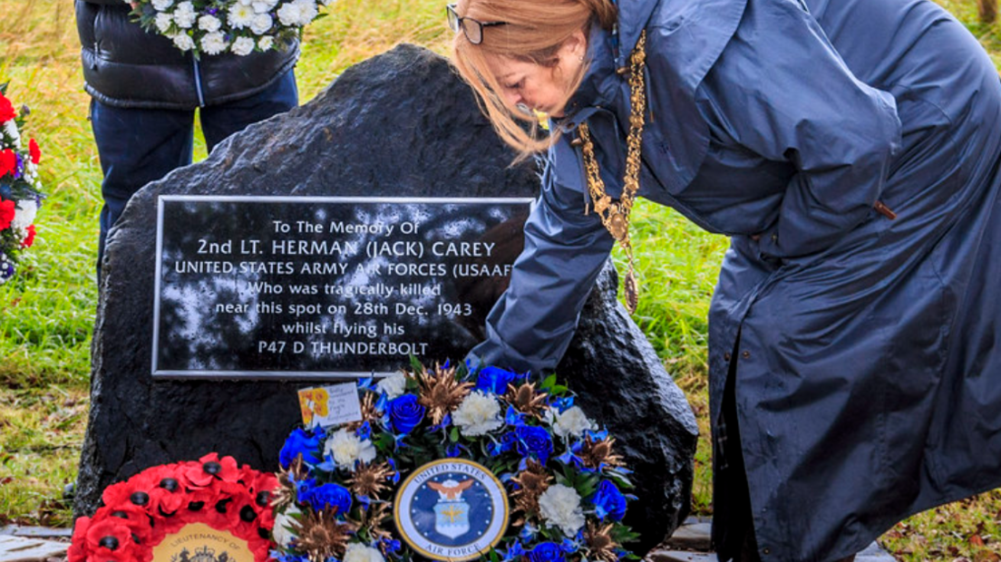 A woman wearing a dark blue raincoat lays a wreath of blue and white flowers next to a wreath of red poppies at the memorial. The memorial is a black slab of stone with a plaque with the pilot's name and details.