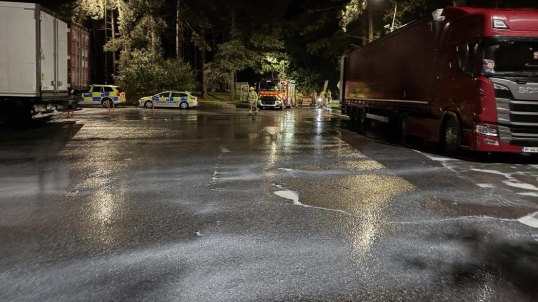 A lorry car park at a service station. The tarmac is covered in a layer of milk. In the background, police cars and a fire truck can be seen.