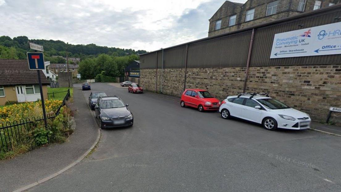 A residential/industrial street lined with parked cars. On one side appears to be a business premises, with homes on the other. Leafy trees are in the far background.