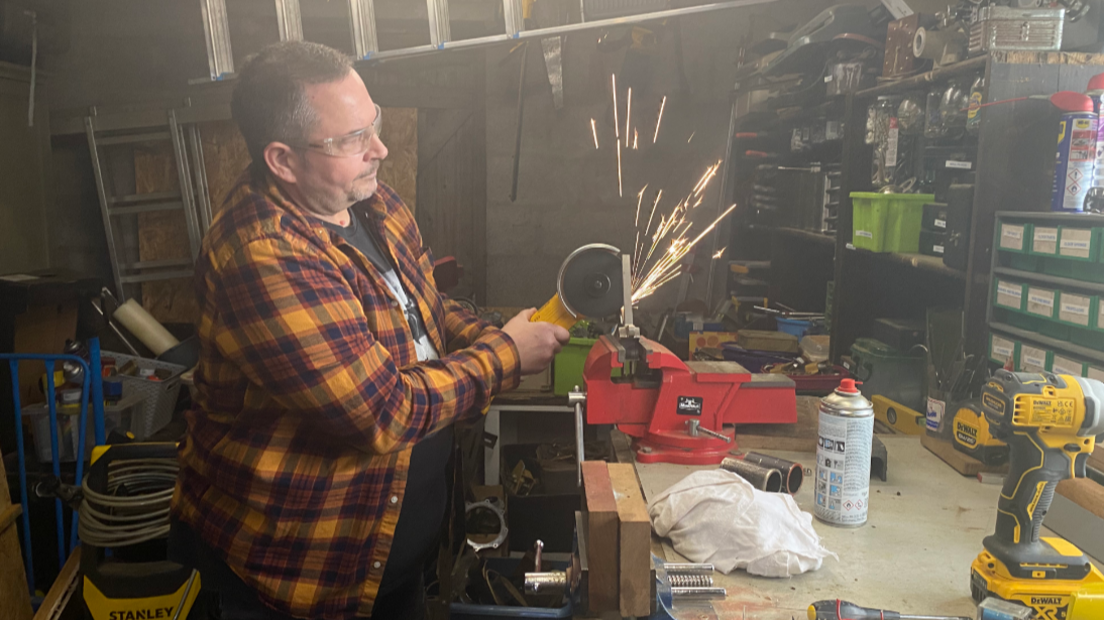 A man in a checked shirt uses an angle grinder to shape a piece of metal in a vice. He is wearing safety glasses and sparks are flying up from the metal