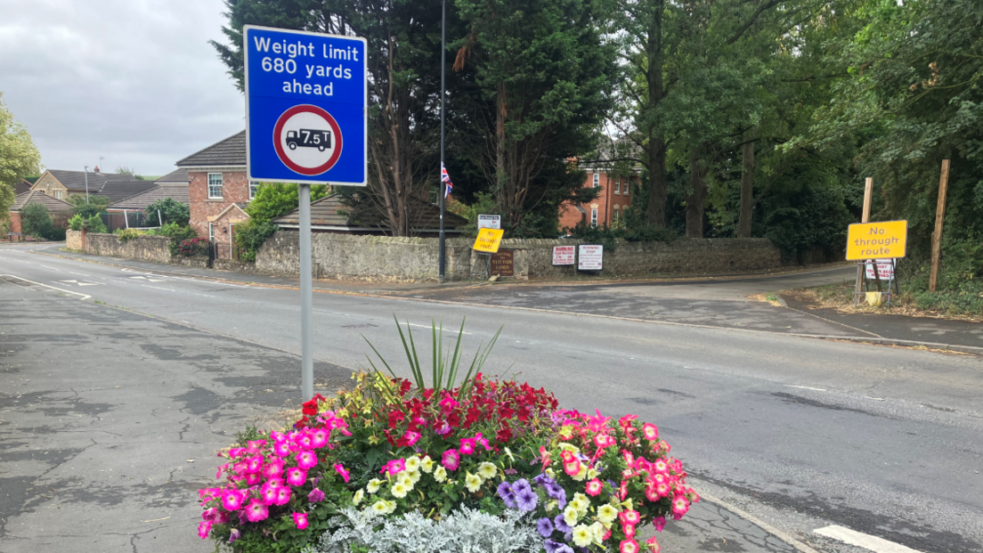 A blue highways sign reads "Weight limit, 680 yards ahead". An image of a lorry is circled in red. In the foreground sits an idyllic village planter, full of coloured flowers. In the background, signs read "No through route".