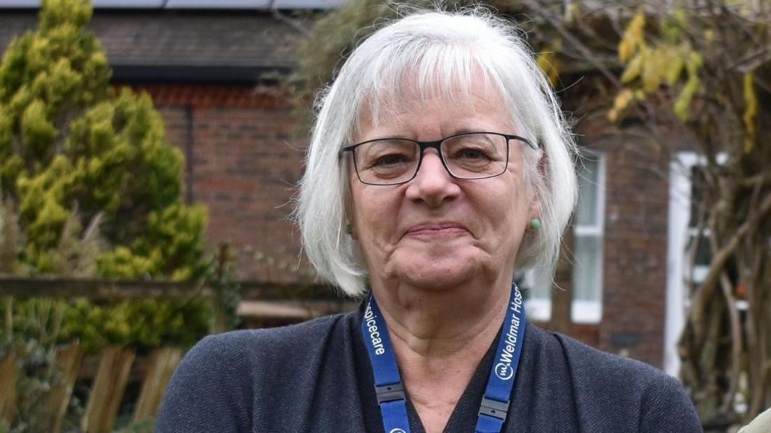 Caroline Hamblett smiles at the camera in a garden at the hospice. She has short, white hair and glasses.