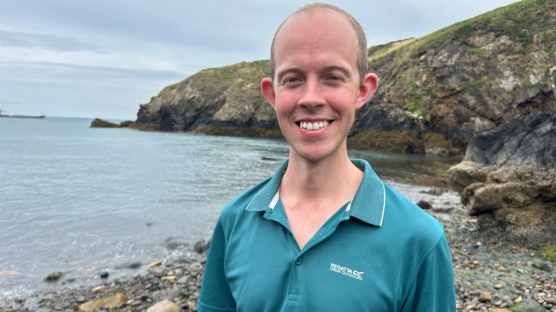 A man in a green coloured polo shirt is standing on a rocky shore. He is smiling at the camera, and behind him is a coastal headland, with sea to his left