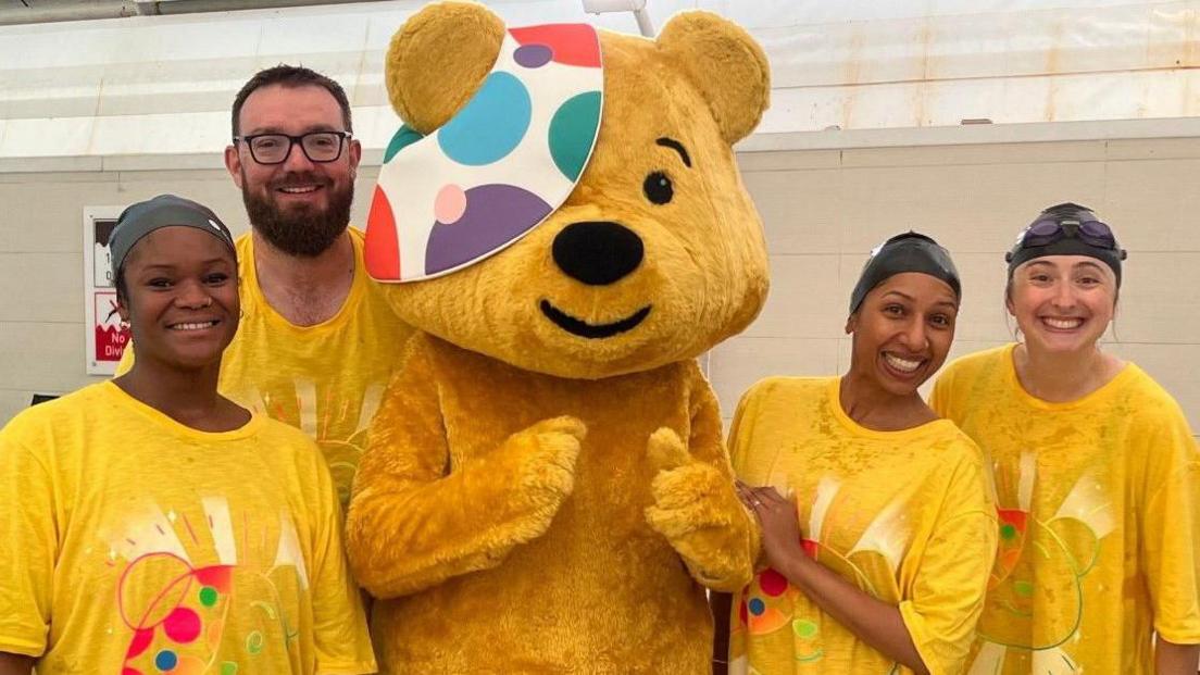 Sali Shobowale, Phil Mercer, Rena Annobil and Katherine Bett gathered around a giant Pudsey Bear at the pool. They are smiling and posing for the camera, wearing yellow t-shirts and swimming caps.