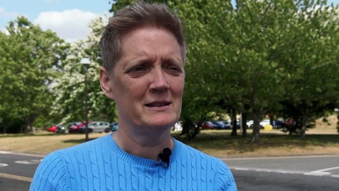 Det Supt Kerry Webb has short cropped grey-blonde hair and is wearing a short-sleeved blue-knit top. She is standing in a street, with a lawned area in the background on which tall and wide trees are standing.