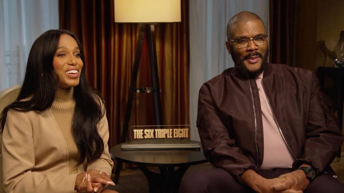 Two people sit on chairs with a table in the middle that has a little sign reading 'The Six Triple Eight' in capitals. Behind them are brown curtains and a tall floor lamp with a white shade. On the left sits a woman with long black straight hair. She is wearing a caramel coloured jumper and she has her hands clasped together, and is smiling. The man next to her has a shaved head, black beard and moustache and wears silver framed glasses. He is wearing a dark coloured bomber jacket over a light coloured shirt. 