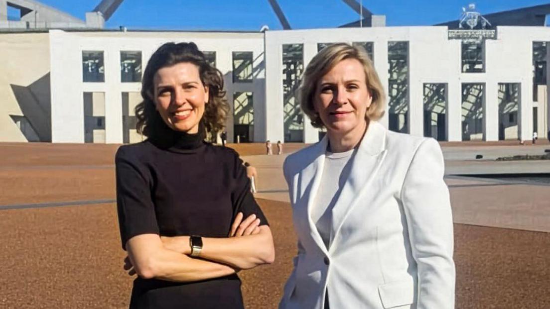Two women standing outside federal parliament house in Canberra. One is a brunette wearing a black top and with her hands folded. The other has a short blonde bob and wearing a white blazer.