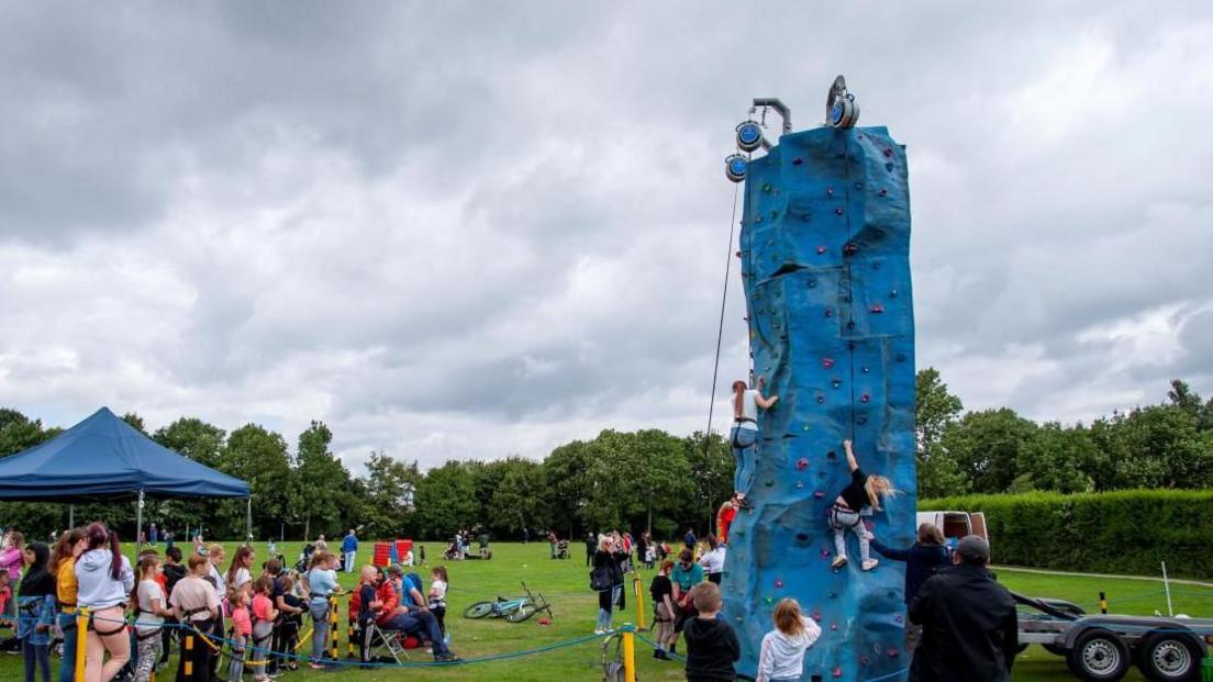 A large blue climbing tower with various young people attempting to climb it in front of a small crowd under a blue gazebo with other children waiting their turn