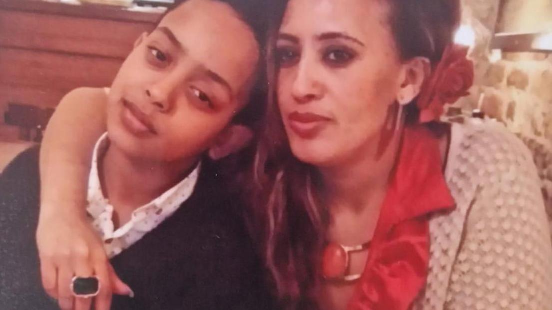 Picture of Biruk on the left, wearing a dark top, leaning into his mum who is wearing a light colour cardigan and red top. She has a red rose in tucked above her ear and has her arm around her son.