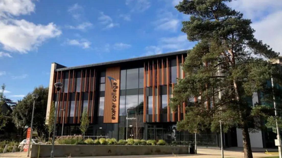 A picture of the front of Exeter College. There is a number of trees and hedges in front of the building with is made out of glass and wood.