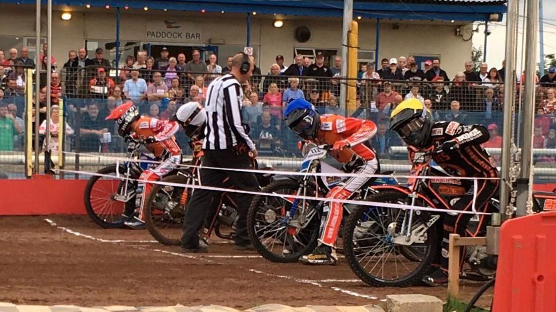 Speedway riders on motorbikes lined up behind the starting line, with a referee in the middle and a crowd of people in the background