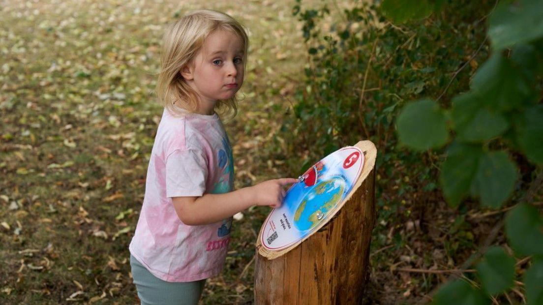 A child pointing at a sign for a book trail on a tree stump. She has on a pink T-shirt and has long fair hair. Leaves are behind her and she is by a tree.