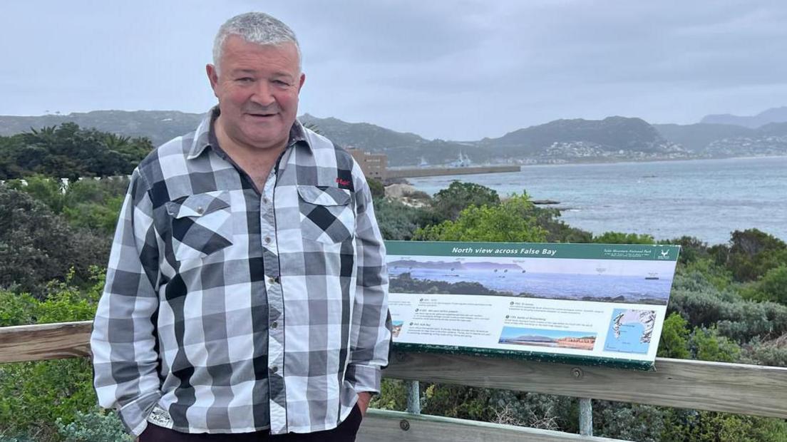 Ivor Wallace - a white/grey haired man wearing a gingham black and white shirt standing in front of a sea bay. 