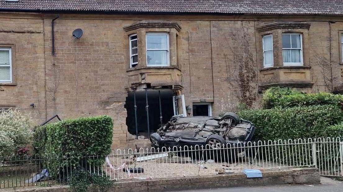 The exterior of the home where the car crashed. A car is on its roof in the front garden, which is surrounded by metal fencing. There is a large hole in the front of the building and it is being held up by scaffolding.