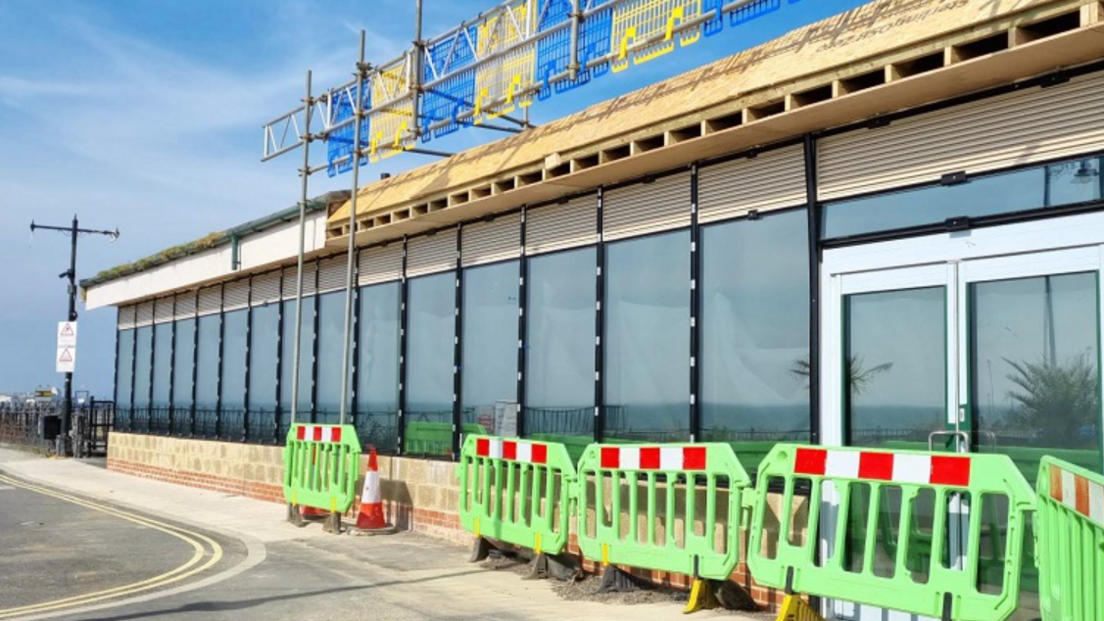 A one-storey building made up of glass windows. In front of the building are green building work barriers.