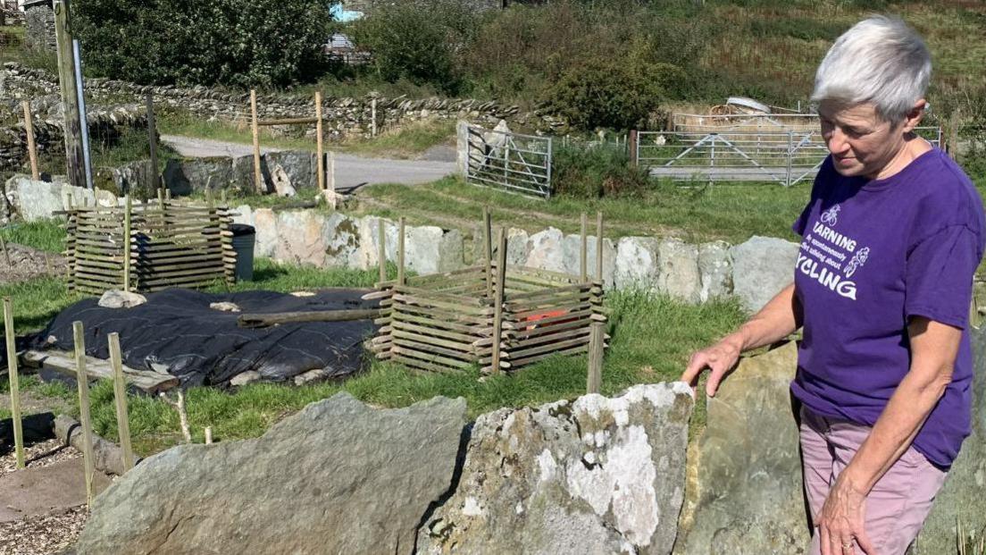 Farmer Celia Caulcott stands next to shard interlocking fencing in farm land. The fencing is roughly shaped and has lichen growing on it. Celia has short great hair and is wearing a purple t-shirt with pink trousers.