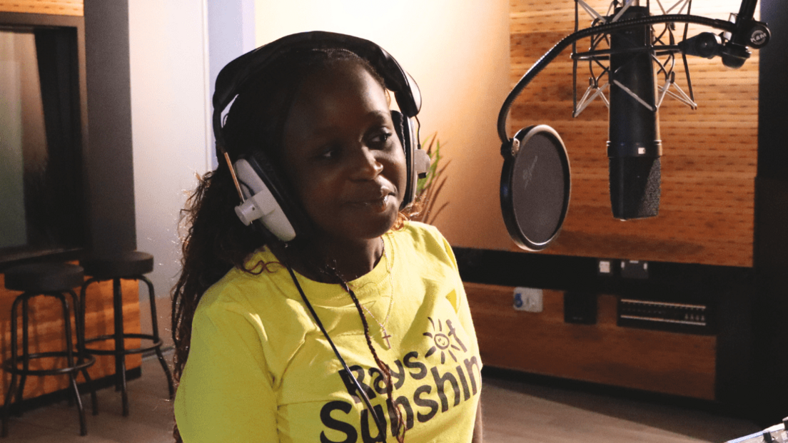 Ruth has long dark hair and is wearing a yellow t-shirt. She is in a recording studio and is wearing headphones whilst stood in front of a microphone