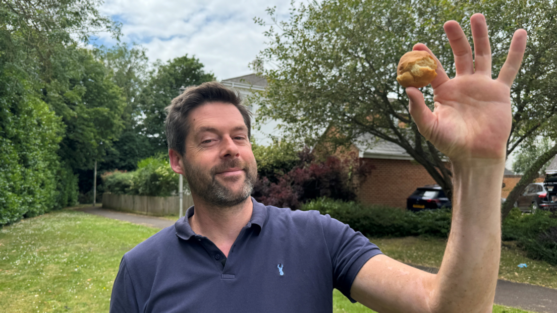 Man holding a scone-like round biscuit in his hand. Looking at the camera. In a small green field. He is wearing a navy top.