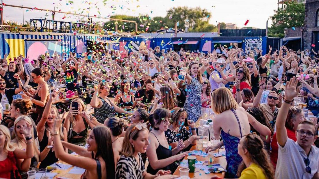 A crowd of people sit at tables outside a court yard on a bright day. People raise their hands as confetti falls from the sky