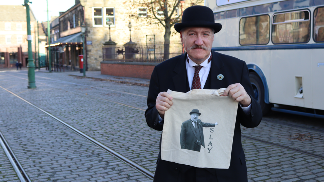 Ian Bean is wearing a historical costume consisting of a bowler hat and a smart black suit. He is holding up a tote bag with his own image on. The text next to him reads: "SLAY".