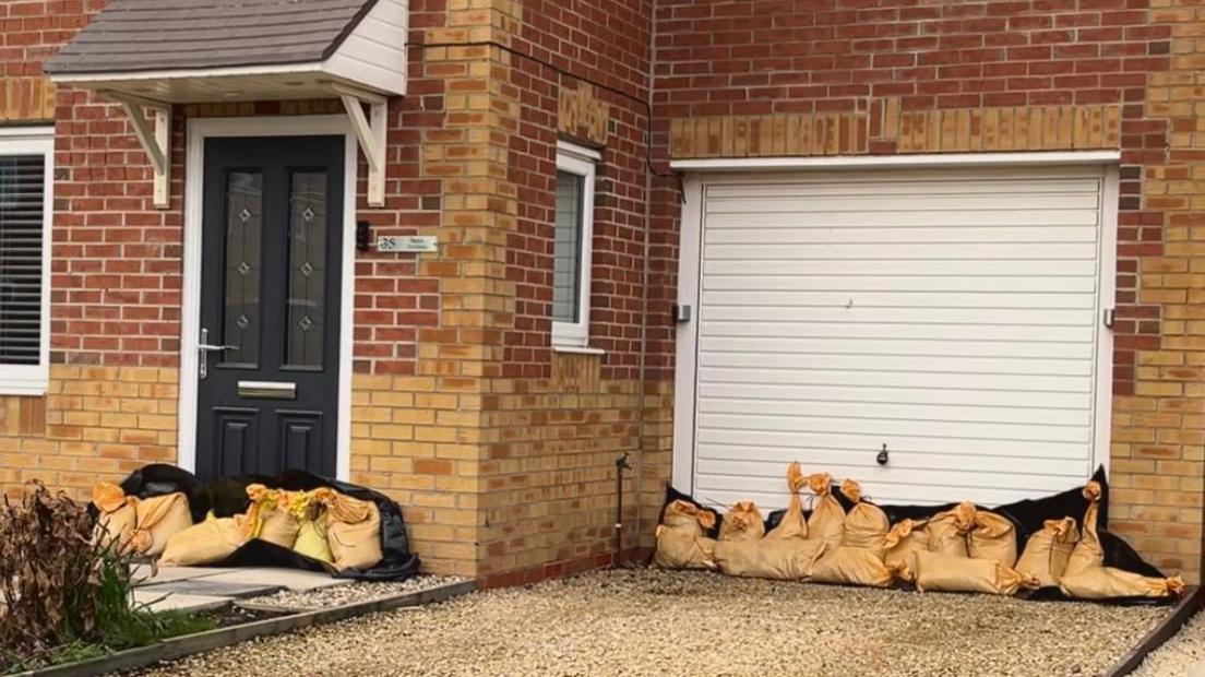 A brick new-build house with a porch and gravel drive way up to a white garage door. There are sandbags propped along the bottom of the front door and the garage door.