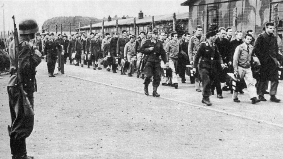 The image shows German soldiers escorting PoWs into the camp, surrounded with barbed wire and huts in the background. In this photo they are US servicemen. It is a black and white image.