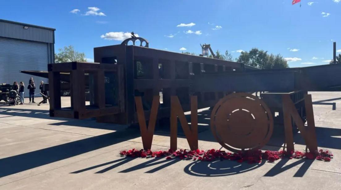Part of the large metal structure lies on the floor with metal letters V N N and a row of poppies underneath.