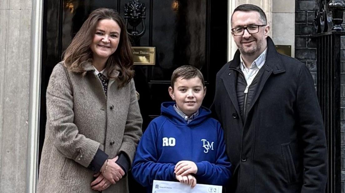 Roman stands in the middle of his dad Ryan and MP Jenny Riddell-Carpenter outside 10 Downing Street. Roman wears a blue hoodie, has short dark hair and holds a letter written on a piece of paper. Ryan has short dark hair, glasses and wears a black coat. Jenny has long dark hair and wears a long grey coat.